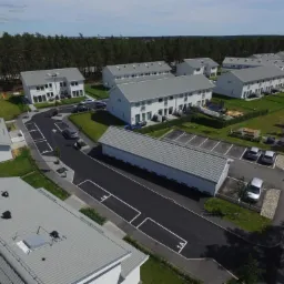 Widok z lotu ptaka na osiedle nowych, jasnych domów z szarymi dachami, otoczonych zielenią i lasem w tle, z widoczną infrastrukturą drogową i parkingową.