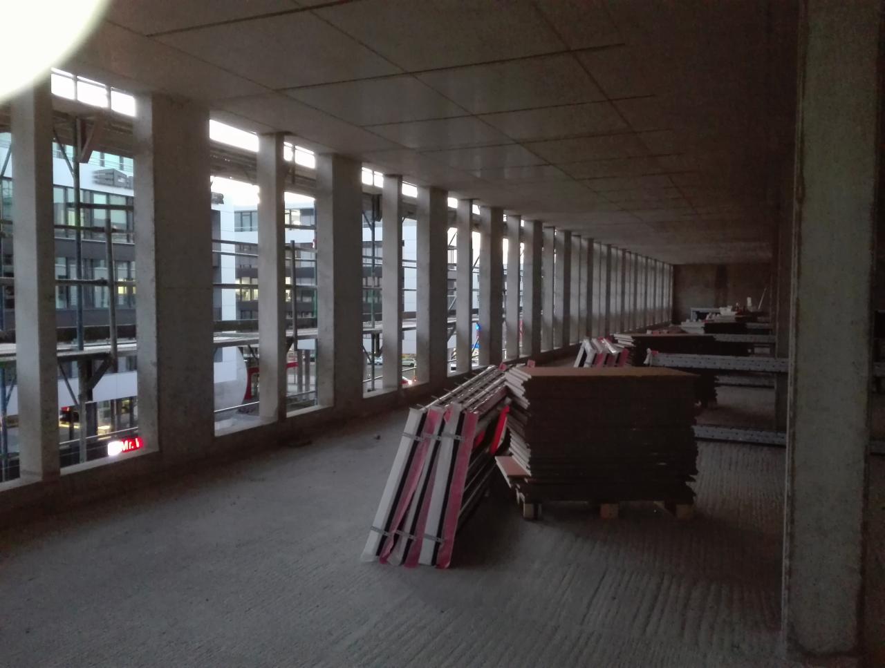 Surowe wnętrze w trakcie budowy z rzędem okien i przygotowanymi panelami, prawdopodobnie do montażu, oświetlone naturalnym światłem z zewnątrz.