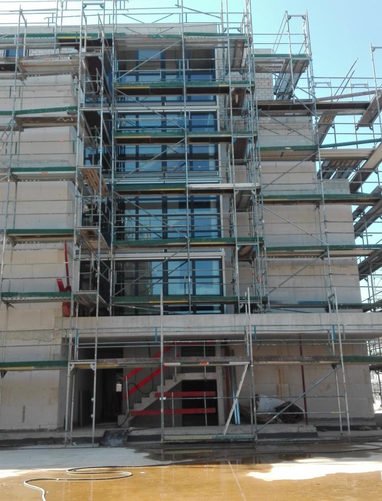 Budynek w trakcie montażu okien, widoczne rusztowania i materiały budowlane, brak wykończenia elewacji.