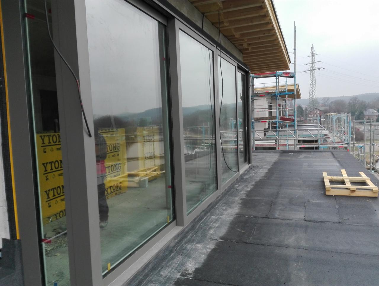 Montaż dużych okien przesuwnych w surowym stanie budynku, widoczne materiały budowlane YTONG i rusztowania na zewnątrz, pochmurny dzień.