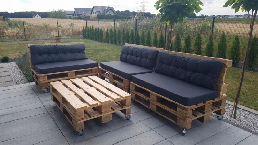 Komplet mebli ogrodowych z palet: sofa, fotel i stolik kawowy z szarymi poduszkami, na kółkach, na tle trawnika i zabudowań.