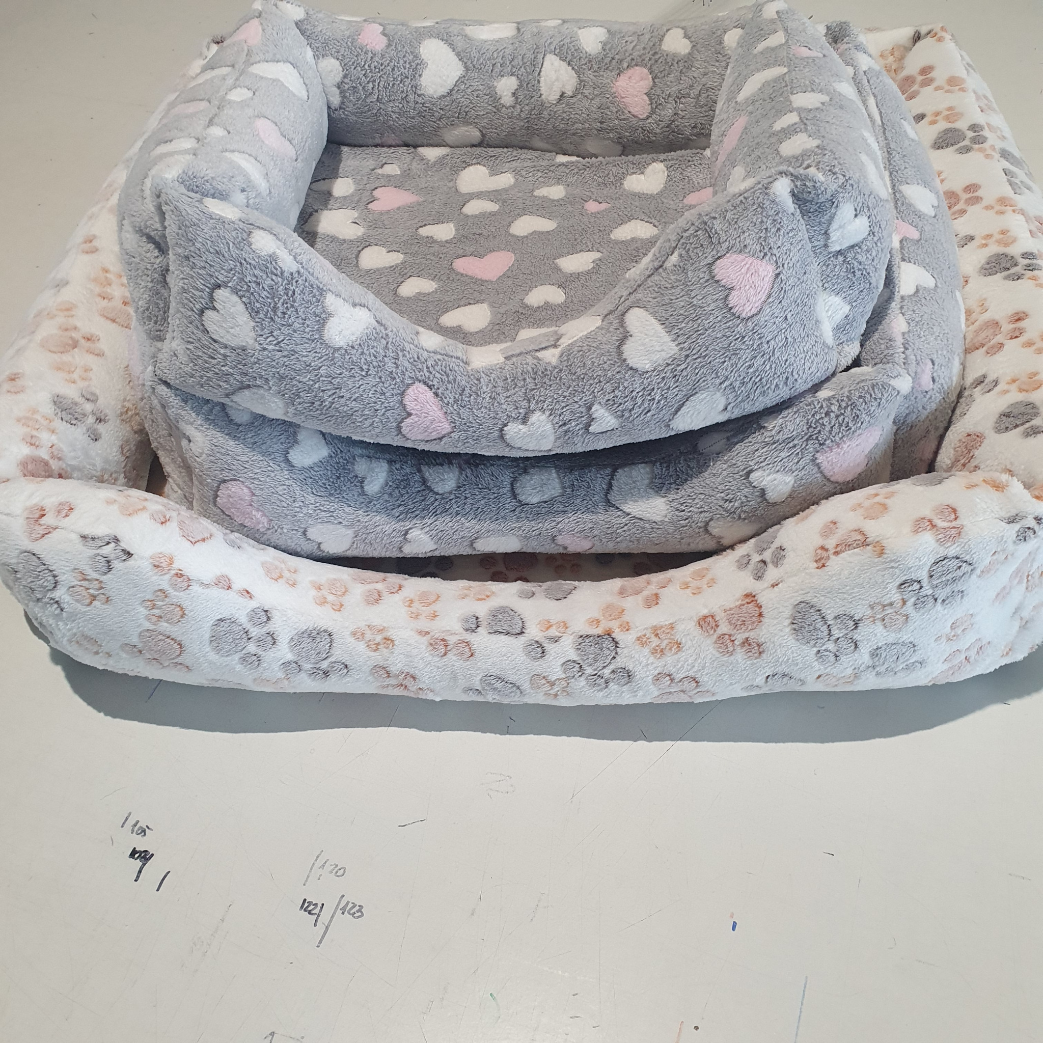 Stos futrzanych legowisk dla zwierząt w odcieniach szarości i bieli z wzorami łap i serc, na białym stole z widocznymi notatkami.