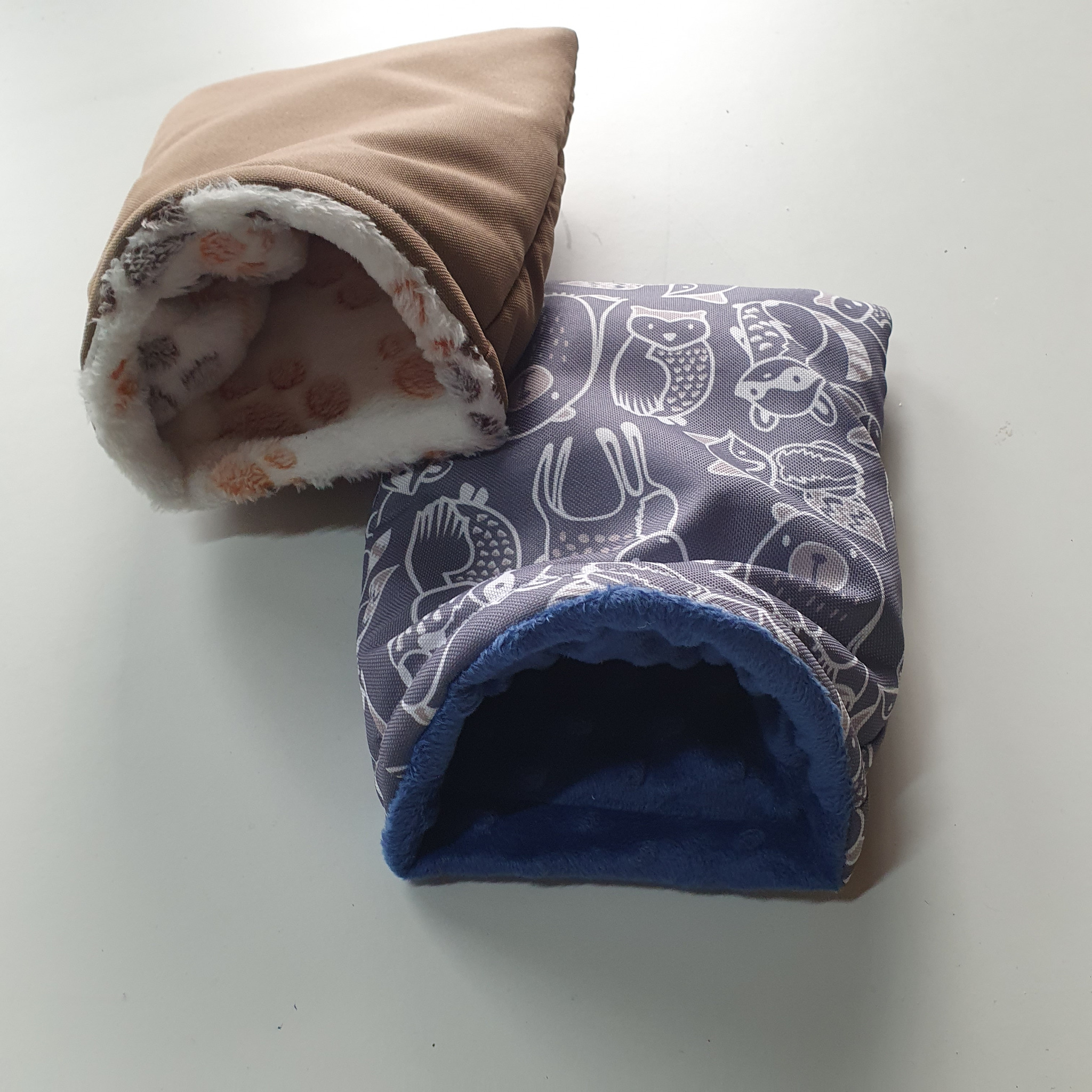 Dwa miękkie, ręcznie robione norki/legowiska dla małych zwierząt: jedno brązowe z futerkową wyściółką w łapki, drugie szaro-białe z motywem zwierzęcym i niebieskim futerkiem w środku.