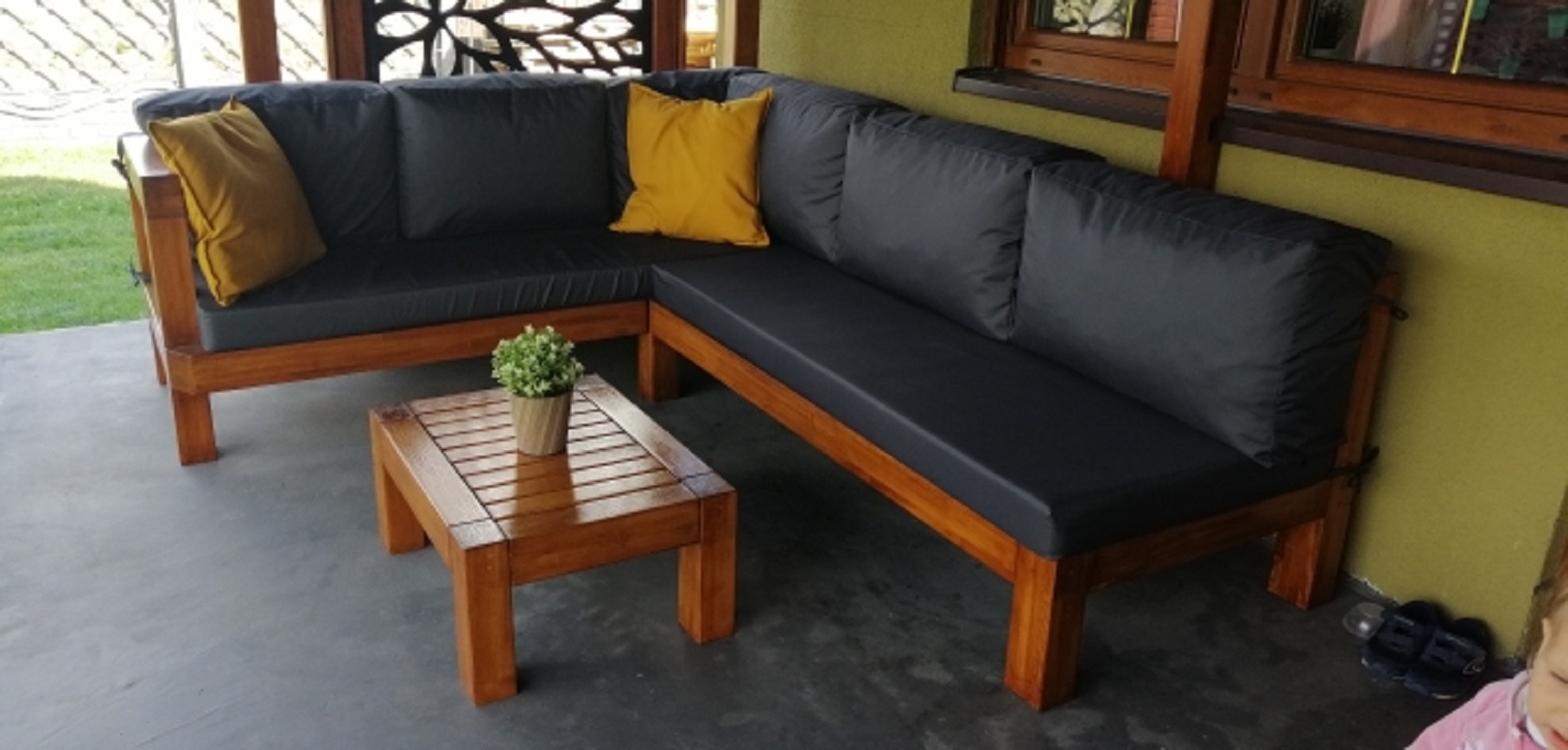 Drewniany narożnik z szarymi poduchami i żółtymi poduszkami dekoracyjnymi na tarasie z drewnianym stolikiem kawowym i rośliną w doniczce.