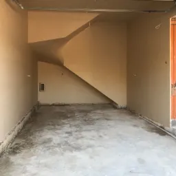 Wnętrze garażu po nałożeniu gładzi gipsowej na ściany i sufit, widoczny surowy betonowy podkład podłogowy, ślady po listwach przypodłogowych i mały otwór wentylacyjny w ścianie.