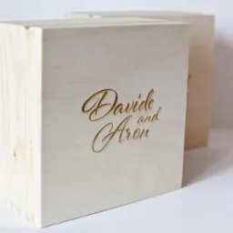 Dwie jasne, drewniane skrzynki z wygrawerowanym laserowo napisem 'Davide and Aron' na froncie, widoczne połączenia na rogach.