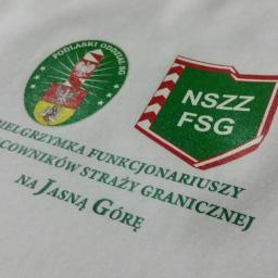 Inter-Gra Artur Szafran - Biała koszulka z zielonymi nadrukami logo i tekstu 'Pielgrzymka Funkcjonariuszy i Pracowników Straży Granicznej na Jasną Górę', widoczny fragment materiału koszulki.