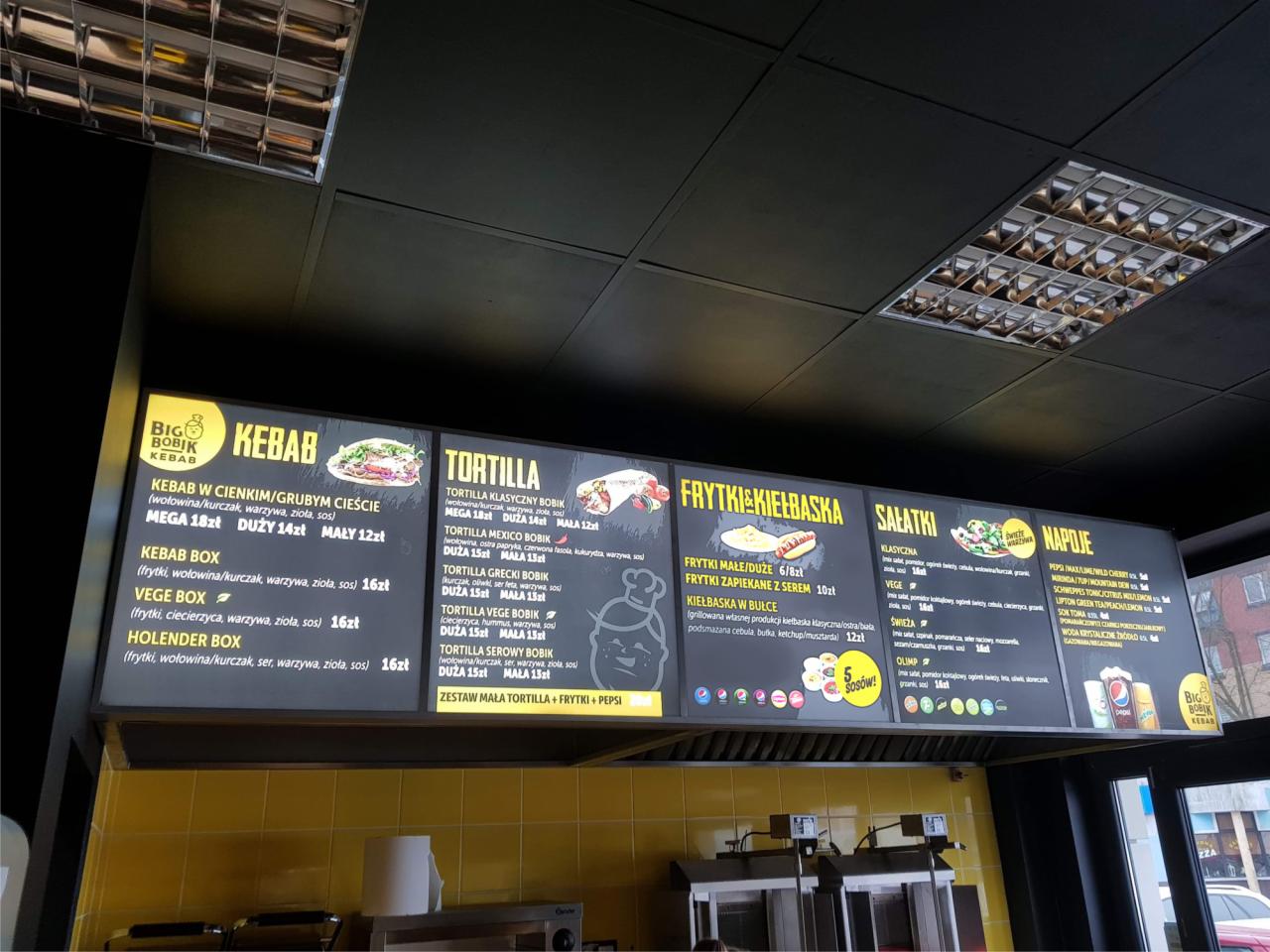 Menu restauracji fast food z kebabami, tortillami, frytkami i napojami, widoczne ceny i składniki.