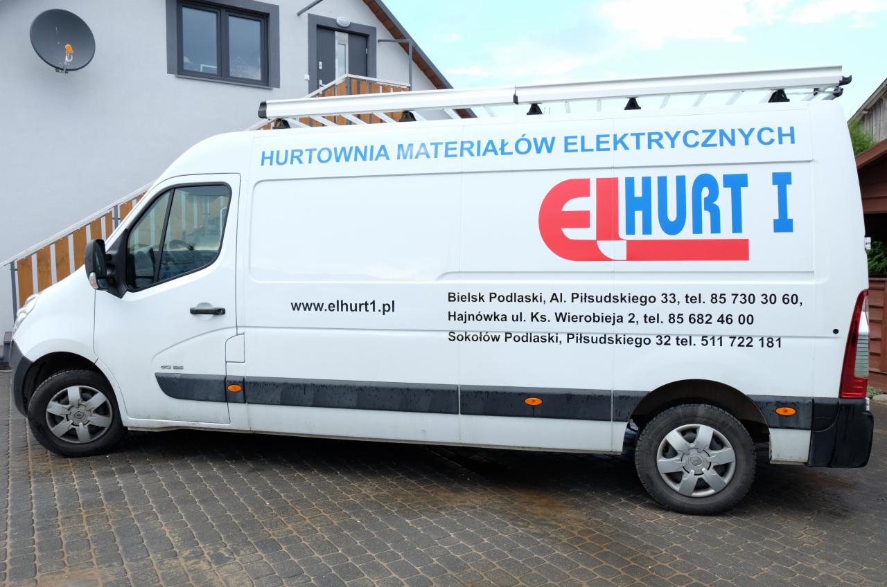 Biały samochód dostawczy z niebiesko-czerwonym logo i danymi kontaktowymi hurtowni materiałów elektrycznych na boku, zaparkowany na brukowanej nawierzchni przed budynkiem.