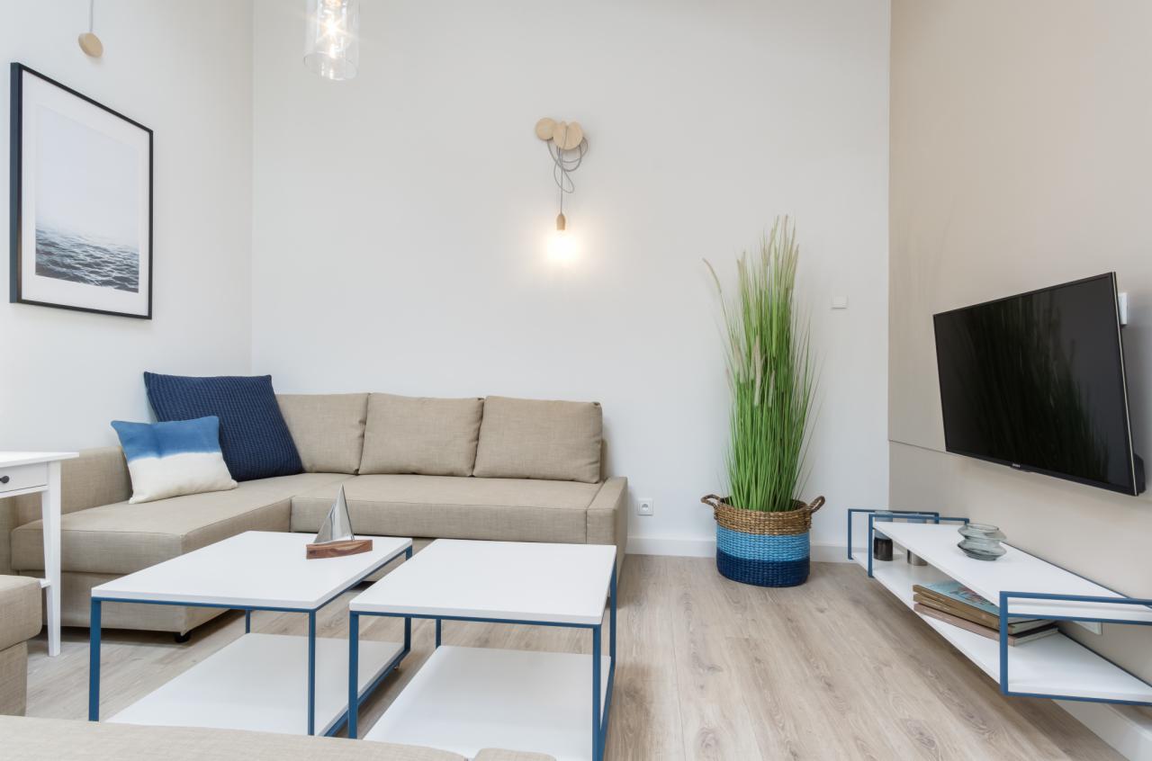 Jasne wnętrze salonu z narożną sofą, dwoma białymi stolikami kawowymi z niebieskimi ramami, telewizorem na ścianie i dekoracjami: lampa wisząca, roślina w koszu i obraz z motywem morza.