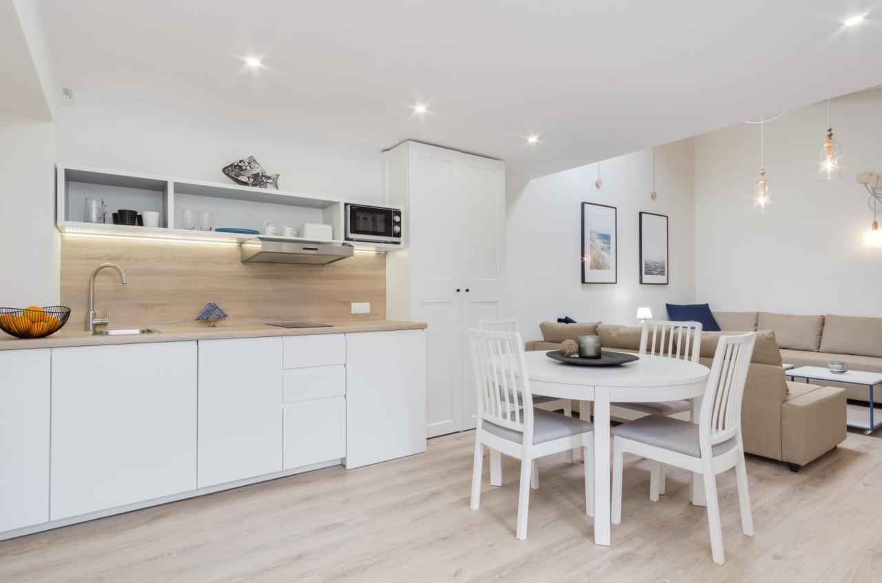 Jasne, nowoczesne wnętrze z aneksem kuchennym, białymi szafkami i drewnianym blatem, okrągły stół z krzesłami oraz beżowa sofa w tle.