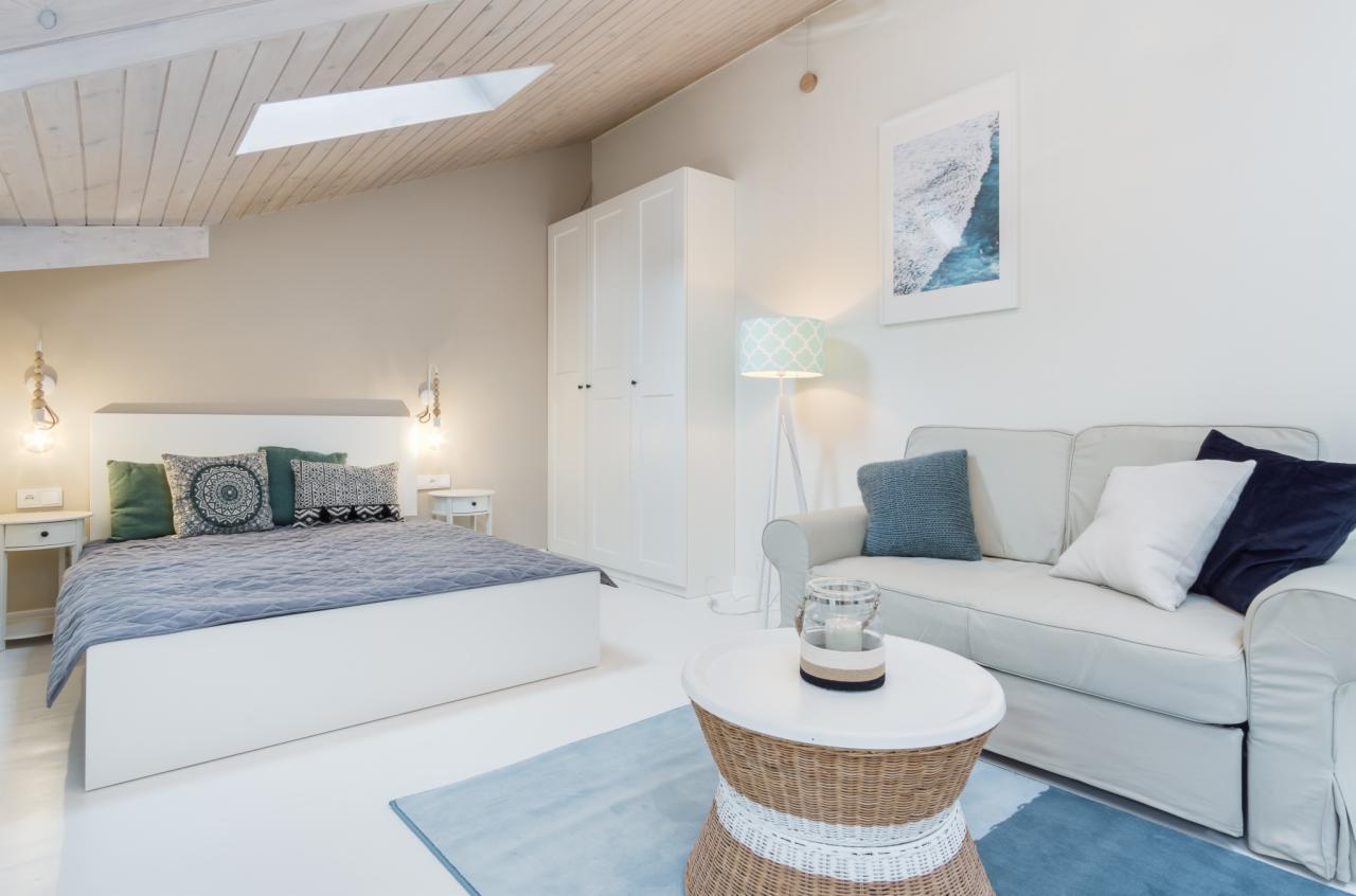 Jasne wnętrze pokoju na poddaszu z drewnianym sufitem, oknem dachowym, białymi ścianami, łóżkiem z szarą narzutą, białą szafą, sofą i okrągłym stolikiem z wiklinowym elementem.