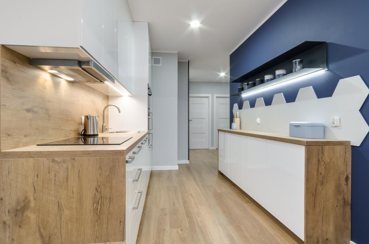Nowoczesna kuchnia z białymi lakierowanymi szafkami, drewnianymi blatami, niebieską ścianą z dekoracyjnymi heksagonami i podświetlaną półką.