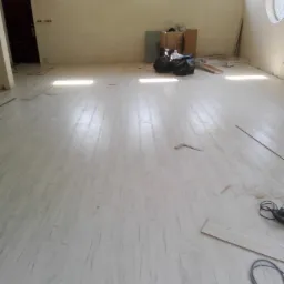 Wnętrze pokoju z ułożonymi panelami podłogowymi o jasnej kolorystyce, widoczne narzędzia pracy i śmieci po montażu, trzy okna w górnej części ściany.