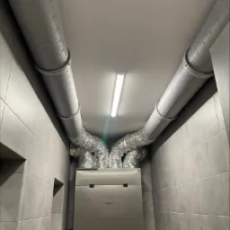 Instalacja systemu wentylacji mechanicznej z odzyskiem ciepła w pomieszczeniu wykończonym szarymi płytkami w Zagrodach. Widoczne rury wentylacyjne i jednostka centralna systemu.