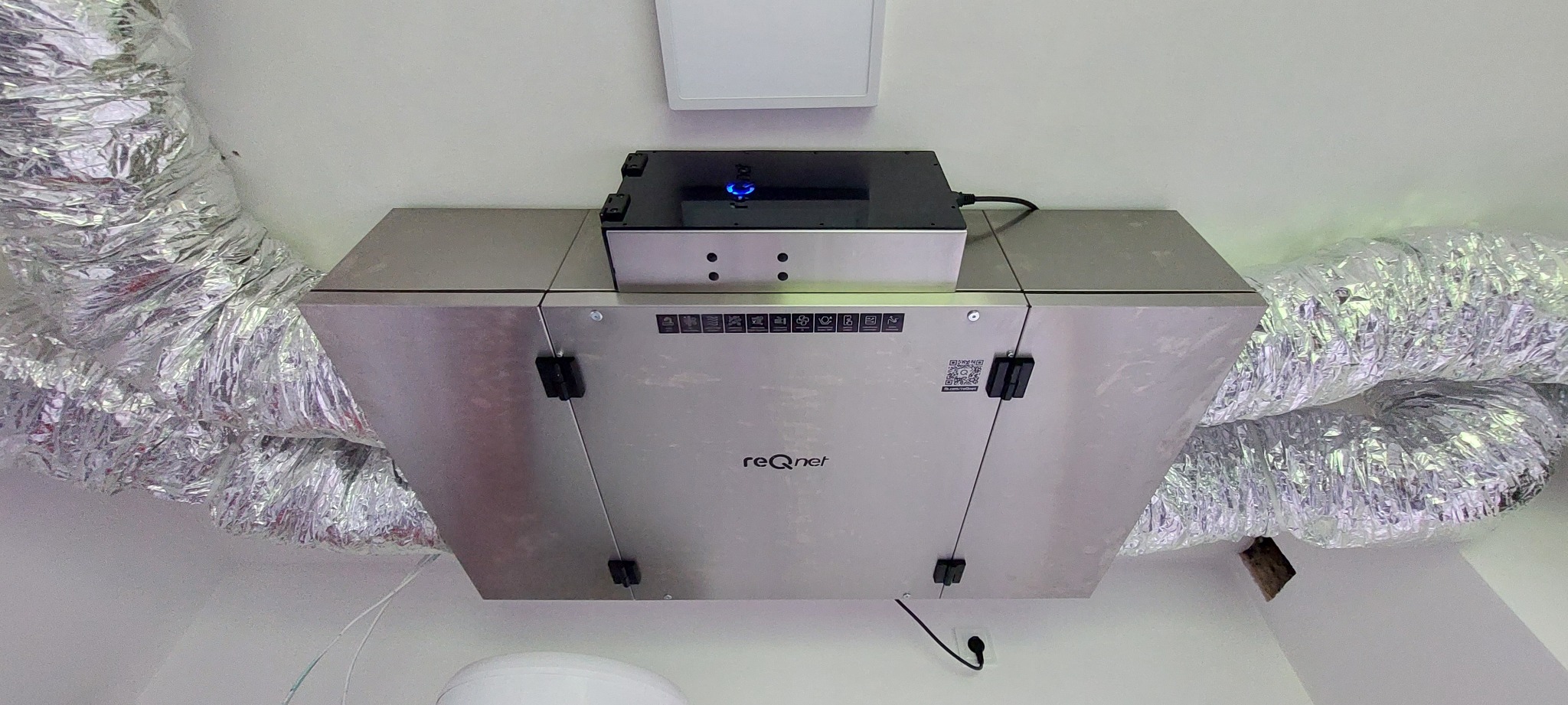 Zamontowana centrala wentylacyjna z aluminiowymi przewodami wentylacyjnymi pod sufitem, widoczny panel sterowania z niebieską diodą i logo firmy na obudowie.