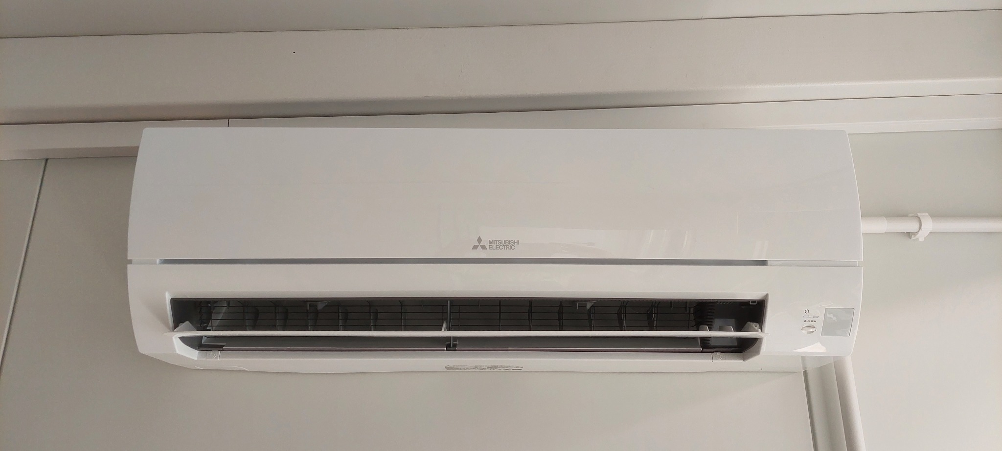 Biała jednostka wewnętrzna klimatyzacji Mitsubishi Electric zamontowana na jasnej ścianie, widoczne rury instalacyjne po prawej stronie.