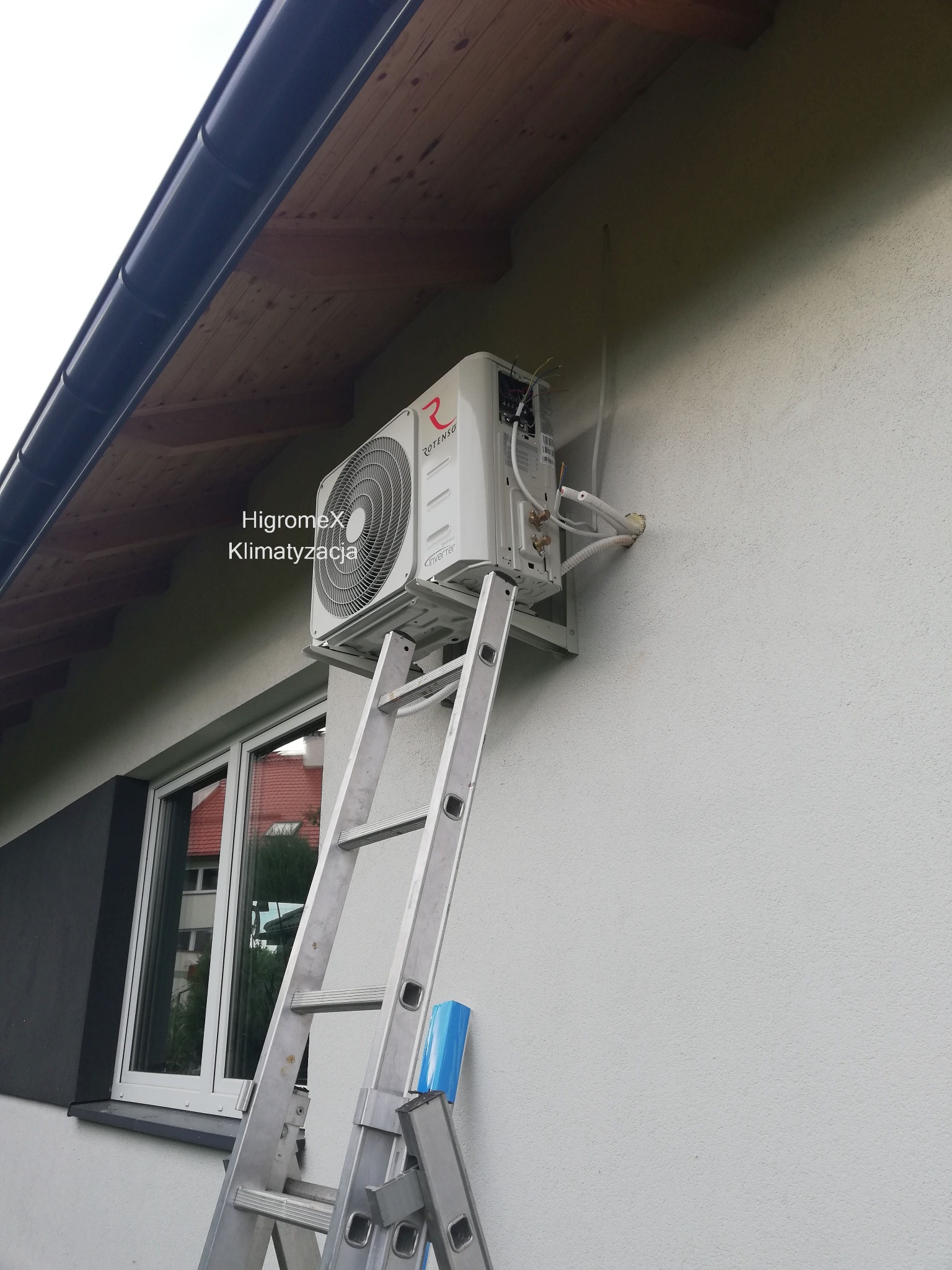 Zewnętrzna jednostka klimatyzacji marki Rotenso zamontowana na ścianie budynku, widoczna drabina aluminiowa i wystające przewody instalacyjne.