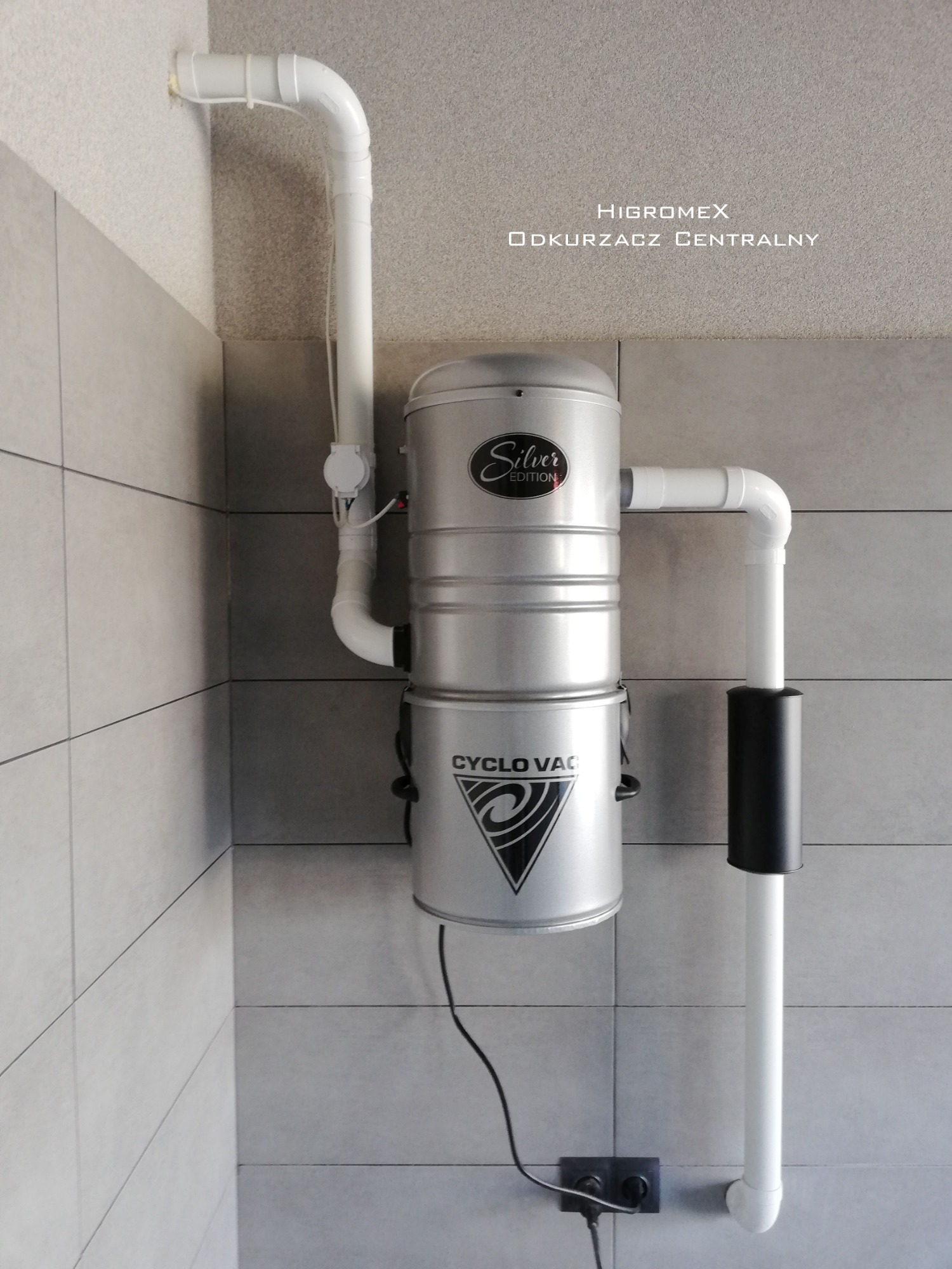 Srebrny, dwuczęściowy odkurzacz centralny marki Higromex, model Cyclo Vac Silver Edition, zamontowany na szarej, kafelkowanej ścianie, z widocznymi rurami instalacyjnymi i gniazdkiem elektrycznym.