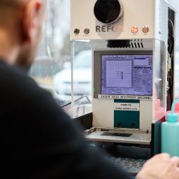Kolego Informatyk - Technik w czarnej koszulce obsługujący maszynę REFOX do laserowego odzyskiwania danych, z ekranem pokazującym siatkę i parametry, obok stoją różowe i niebieska butelka.