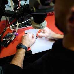 Kolego Informatyk - Technik precyzyjnie manipuluje miniaturowym chipem elektronicznym pod mikroskopem stereoskopowym, używając pęsety i czerwonego narzędzia na białej serwetce. W tle płyta 'MULTIFLASH' i rozłożony...