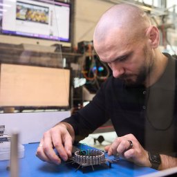 Kolego Informatyk - Mężczyzna w czarnej koszulce precyzyjnie pracuje nad złożonym urządzeniem elektronicznym z okablowaniem, na niebieskiej macie w warsztacie, z monitorami w tle.