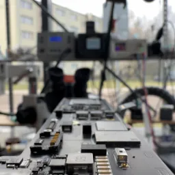 Płyta główna komputera pod mikroskopem, widoczne elementy elektroniczne, sprzęt diagnostyczny w tle, okno z widokiem na budynek.