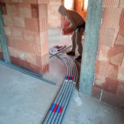 Pracownik w bluzie w paski układa szare rury instalacyjne z czerwono-niebieskimi oznaczeniami w surowym wnętrzu budynku z cegły.