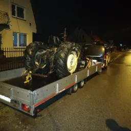 Ciągnik rolniczy zabezpieczony pasami transportowymi na aluminiowej przyczepie typu Temared, podpiętej do ciemnego busa, sfotografowany nocą na tle dom&oacute;w.