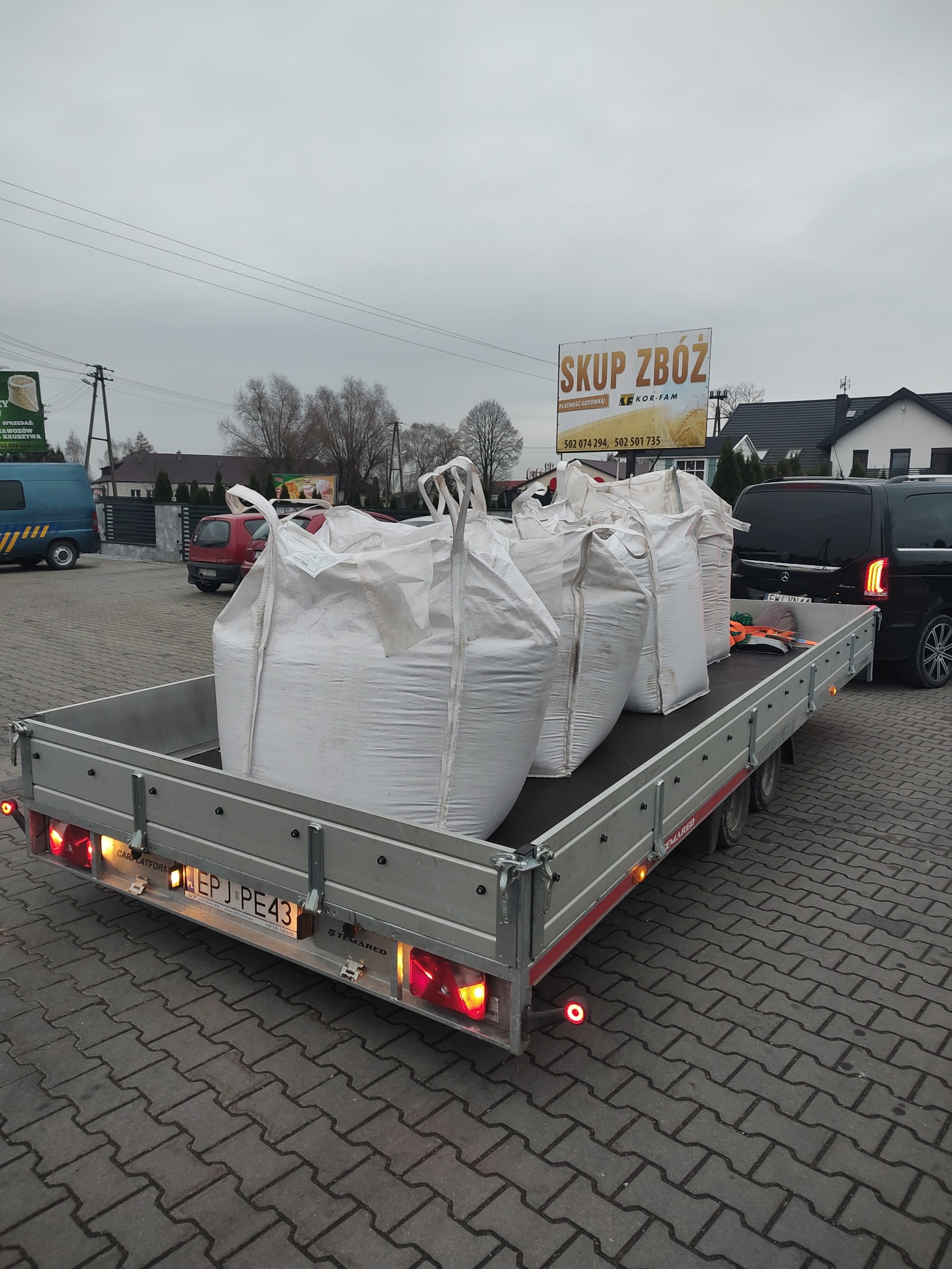 Przyczepa samochodowa z czterema dużymi workami typu Big Bag, prawdopodobnie z ziarnem, zaparkowana na brukowanym placu, w tle budynki i reklama skupu zboża.