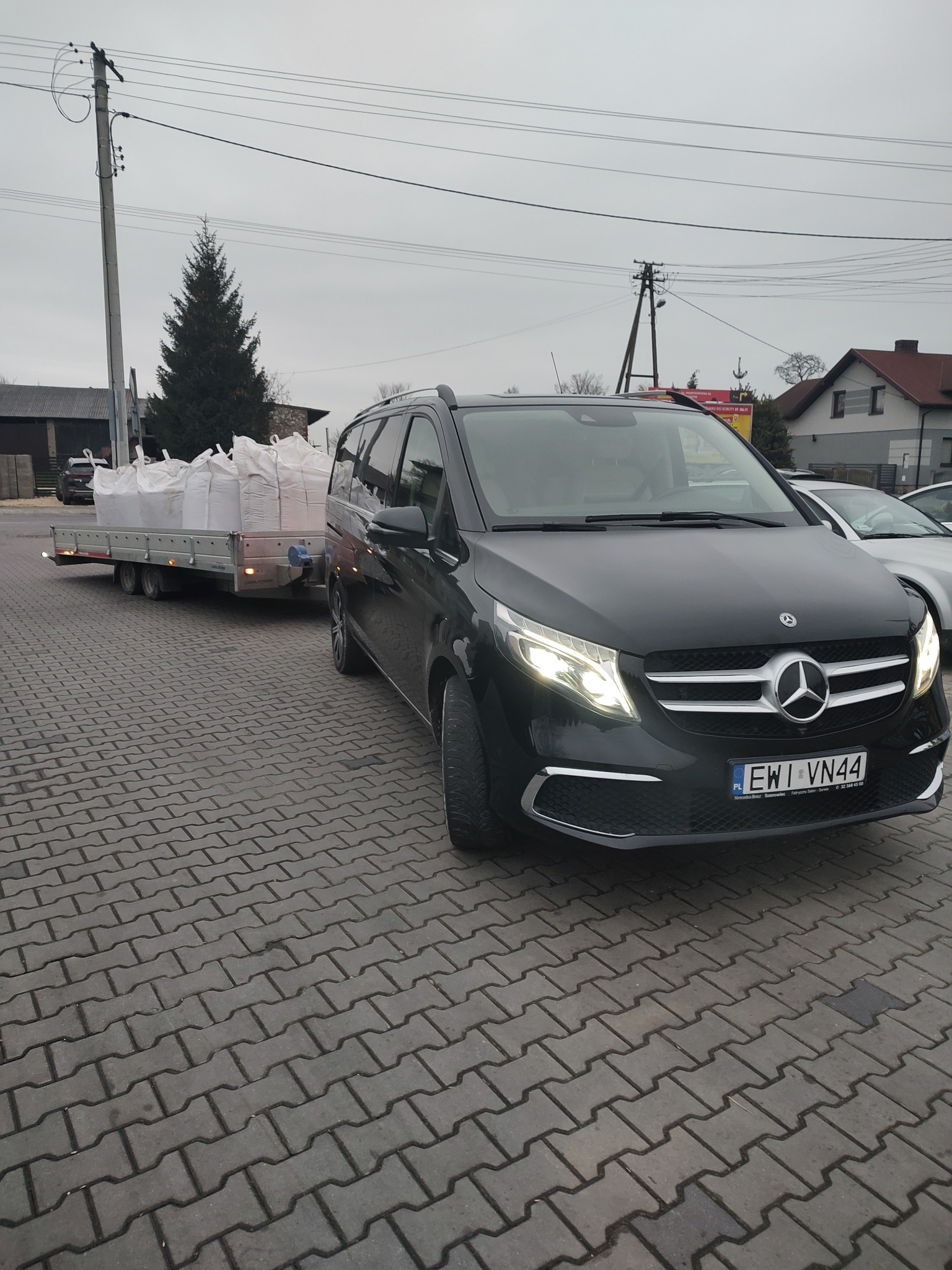 Czarny Mercedes V-klasy z przyczepą wypełnioną białymi workami, zaparkowany na brukowanej powierzchni, z widocznym numerem rejestracyjnym EWI VN44.