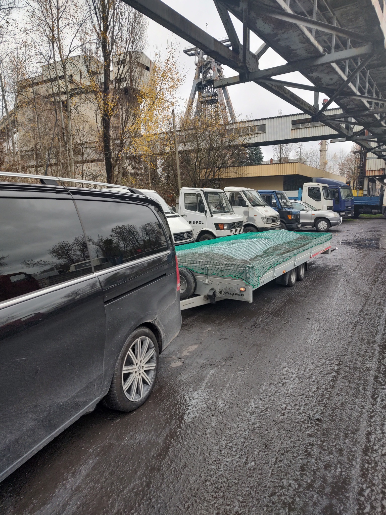 Czarny bus Mercedes z przyczepą lawetą na mokrym placu z widokiem na konstrukcję kopalni w tle.