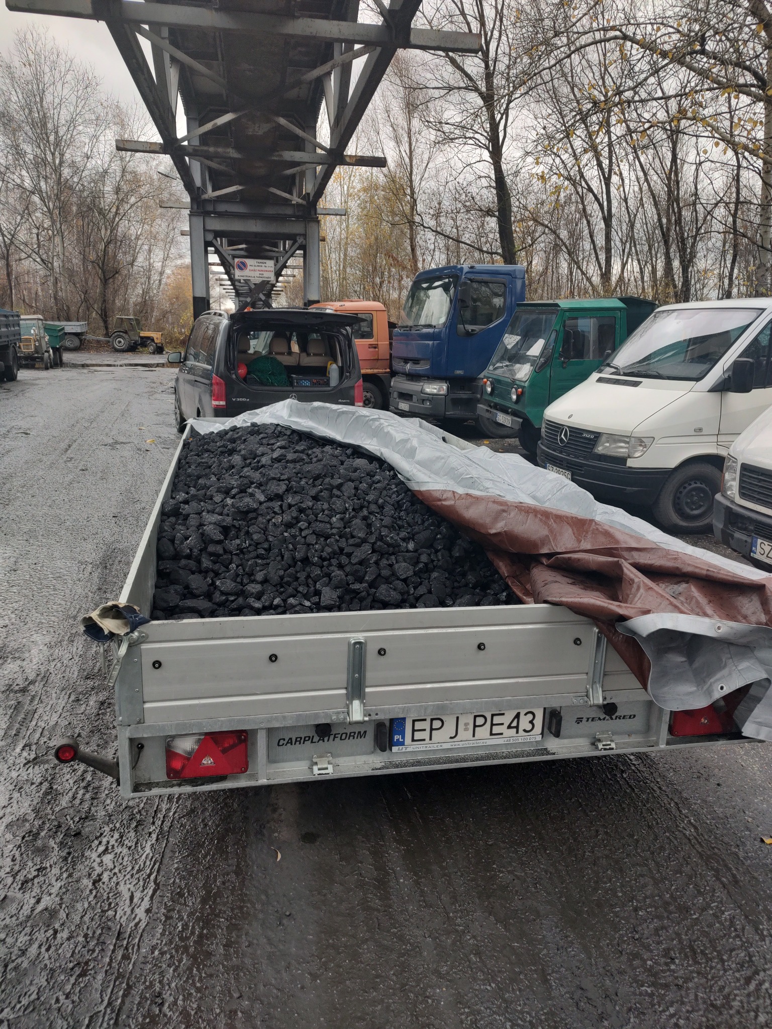 Przyczepa z ładunkiem węgla kamiennego przykryta plandeką, zaparkowana na mokrym placu pod metalową konstrukcją, w tle zaparkowane busy i samochody dostawcze.