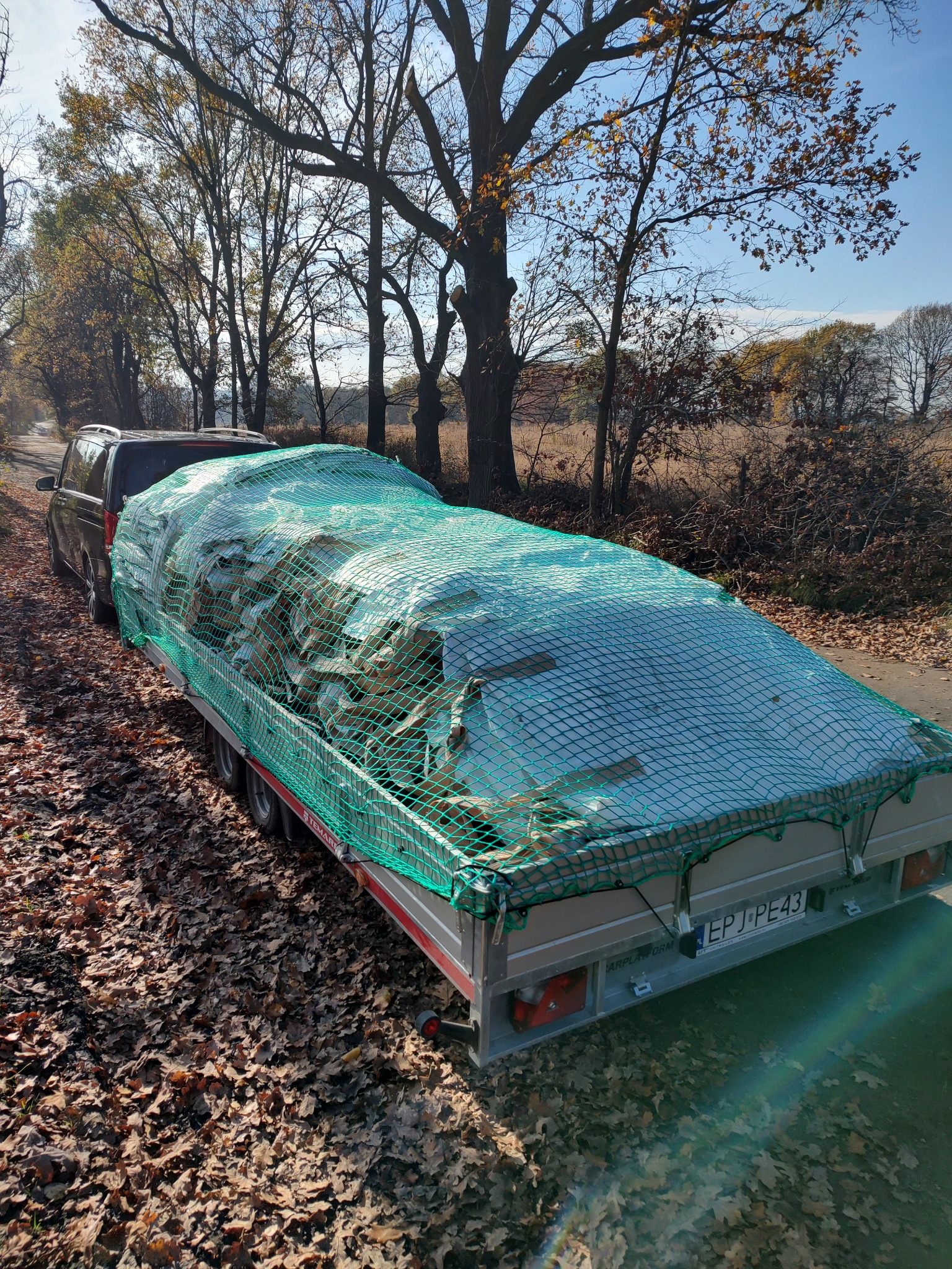 Samochód osobowy ciągnie przyczepę z ładunkiem zabezpieczonym zieloną siatką, zaparkowany na poboczu drogi w otoczeniu drzew z jesiennymi liśćmi.