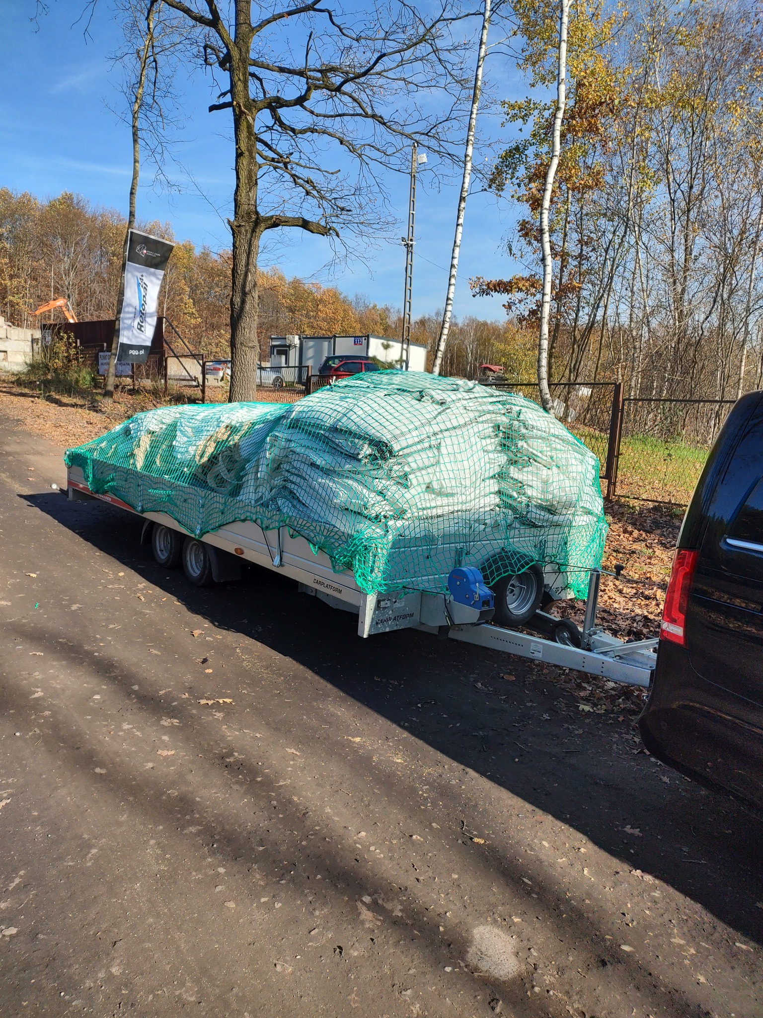 Przyczepa samochodowa z ładunkiem zabezpieczonym zieloną siatką, zaparkowana na poboczu drogi, obok zaparkowany czarny samochód, w tle drzewa i ogrodzenie.