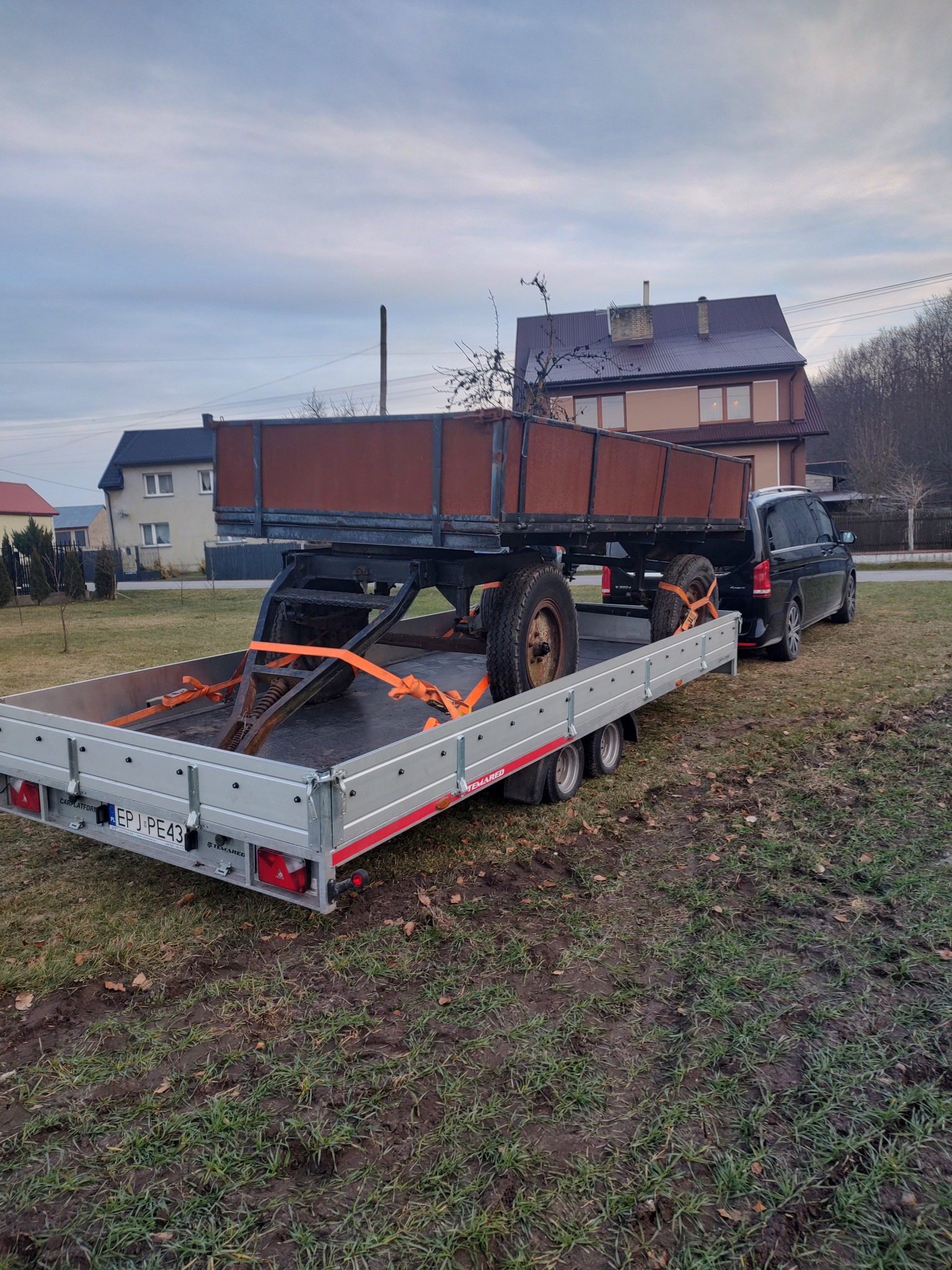 Laweta z platformą transportową i załadowaną przyczepą z kołami, zabezpieczona pomarańczowymi pasami, podłączona do czarnego busa na tle domu z brązowym dachem.