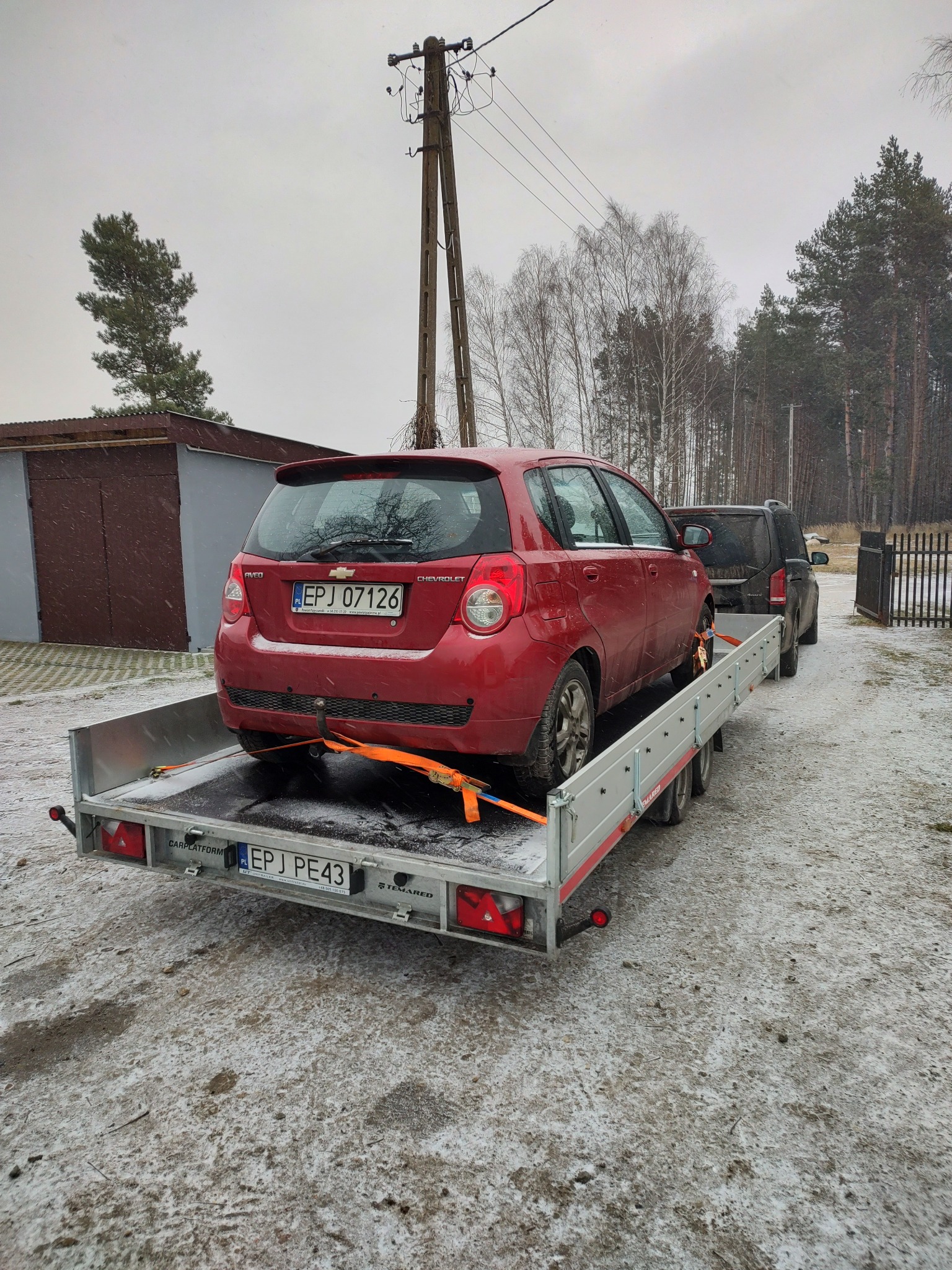 Czerwony Chevrolet Aveo zabezpieczony pomarańczowymi pasami transportowymi na aluminiowej lawecie, widok z tyłu, w tle szary garaż i zaśnieżone drzewa.