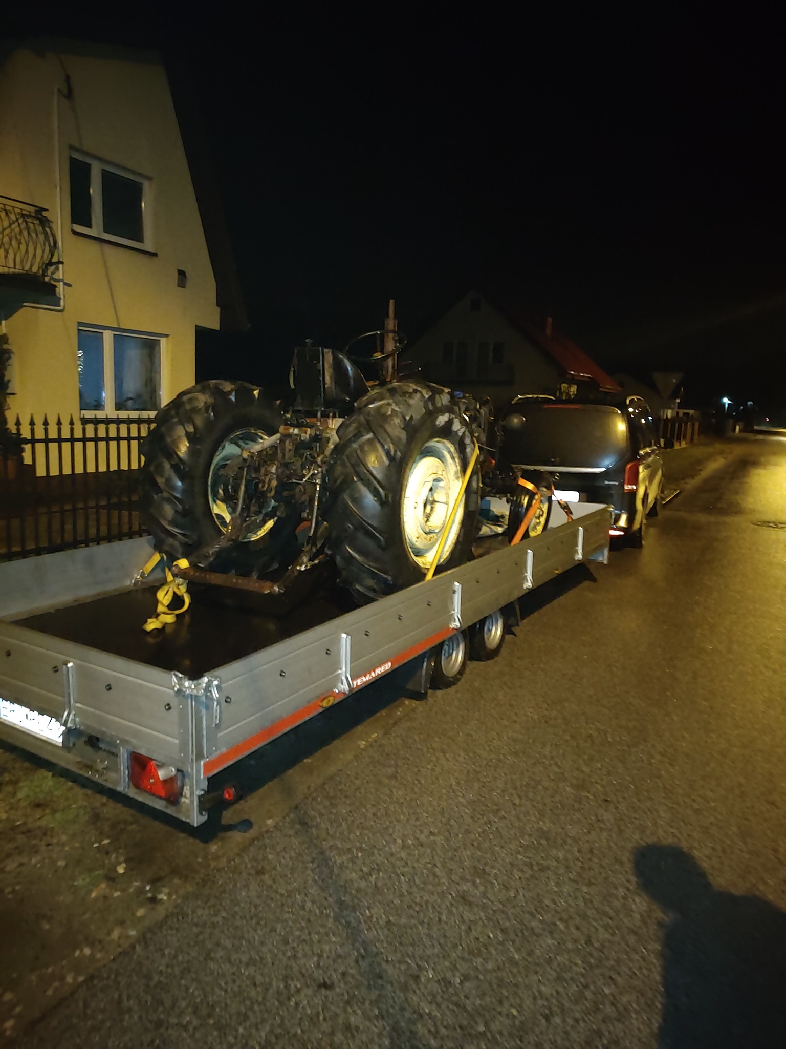 Ciągnik rolniczy zabezpieczony pasami transportowymi na aluminiowej przyczepie typu Temared, podpiętej do ciemnego busa, sfotografowany nocą na tle domów.