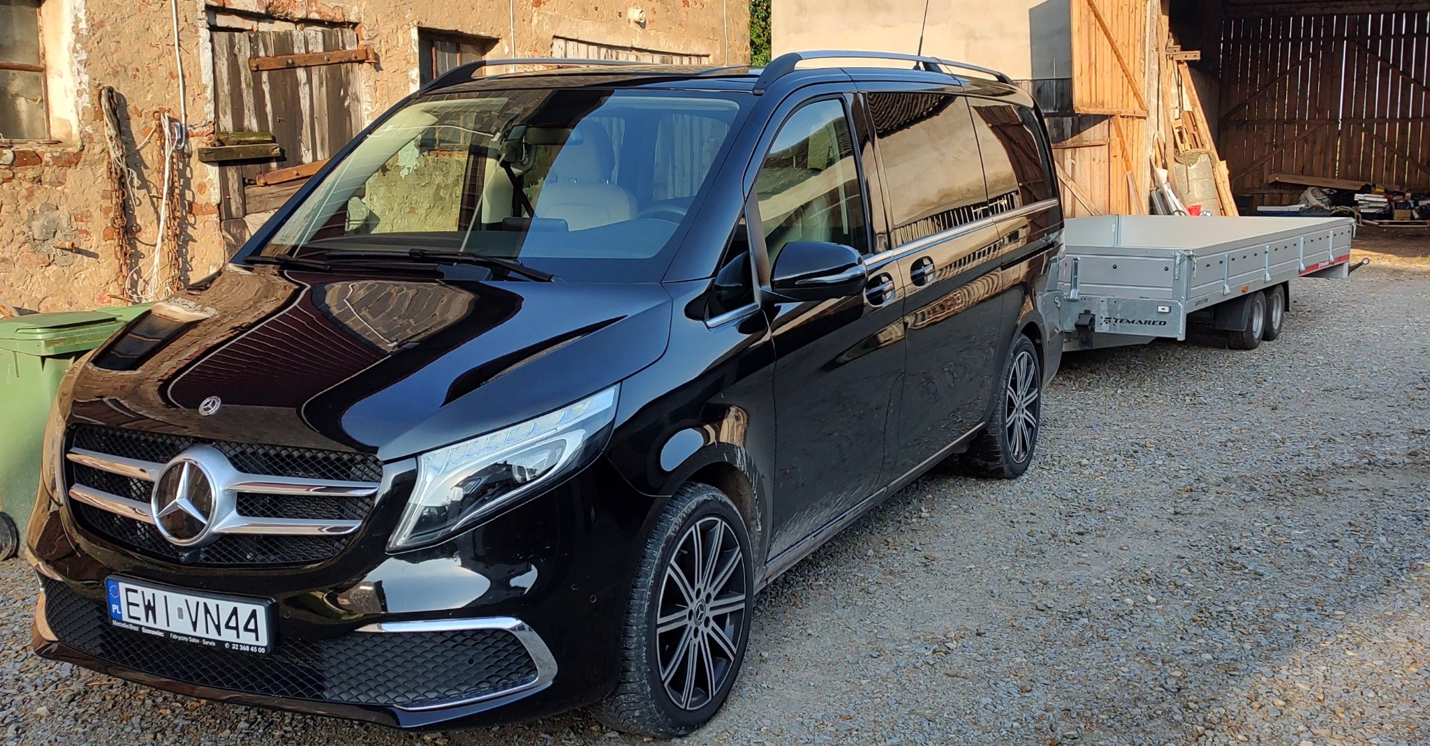 Czarny Mercedes V-klasa z przyczepą platformową marki Temared na żwirowym podwórku, w tle budynek gospodarczy z drewnianymi wrotami.