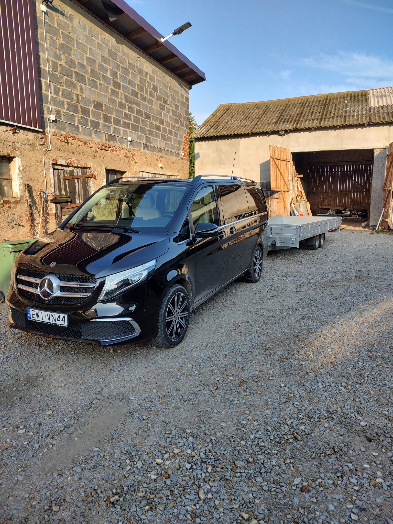 Czarny Mercedes V-klasa z przyczepką na żwirowym podwórku, obok budynków gospodarczych z cegły i drewna.