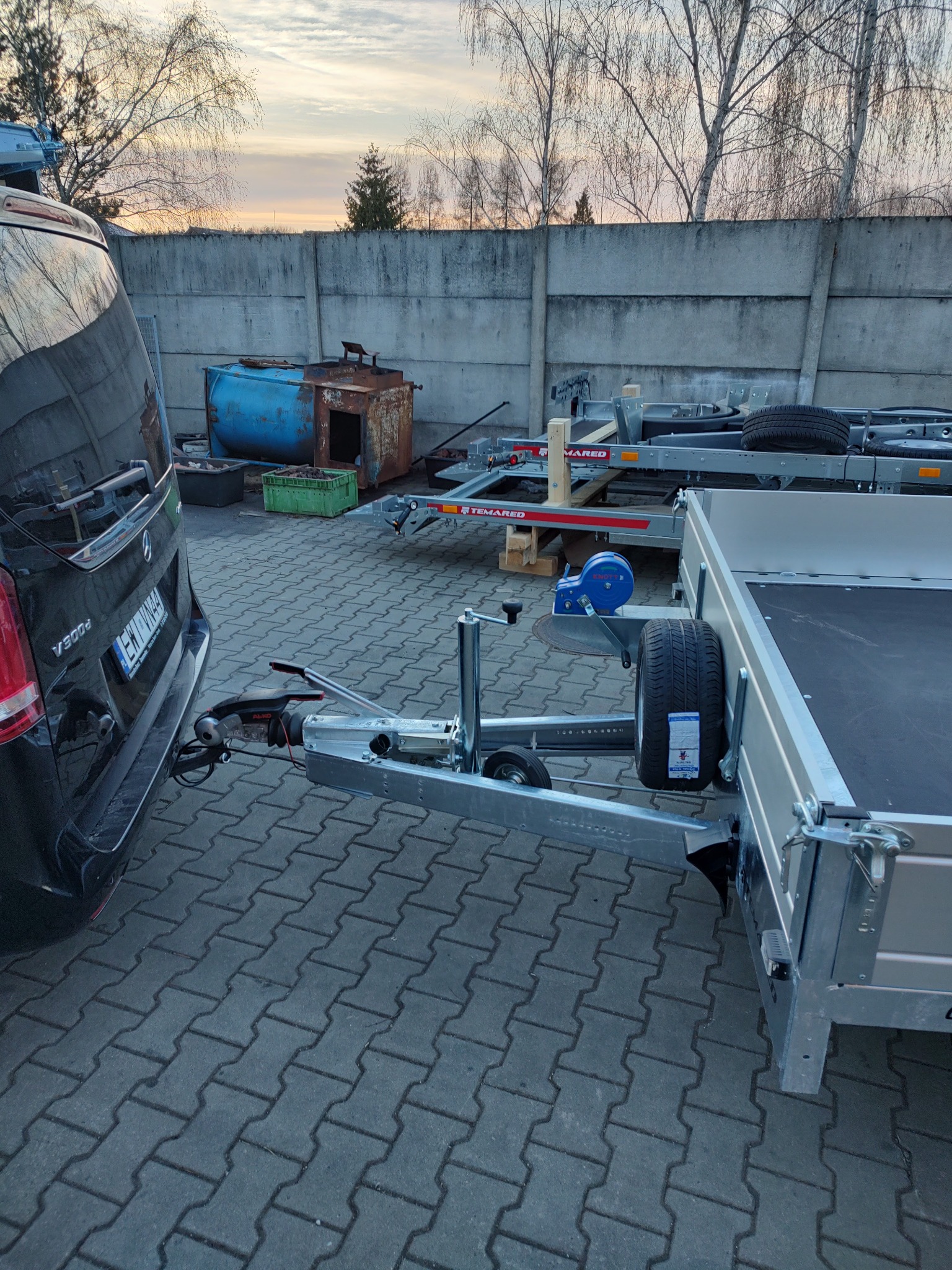 Czarny van marki Mercedes V300d z podpiętą nową, srebrną przyczepą na kostce brukowej, w tle betonowy mur i inne przyczepy.