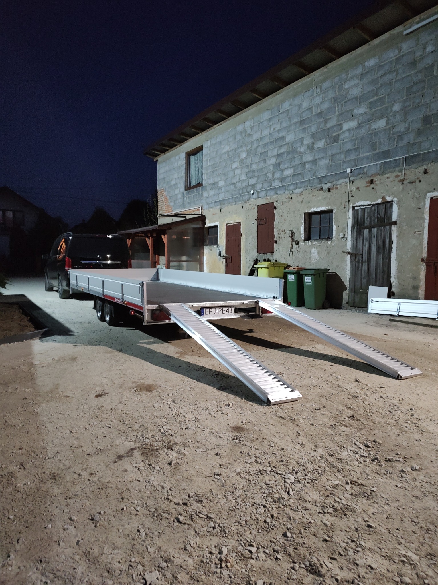 Przyczepa samochodowa z aluminiowymi rampami rozłożonymi do załadunku, zaparkowana na żwirowym podłożu przed budynkiem gospodarczym z cegły w wieczornym oświetleniu.
