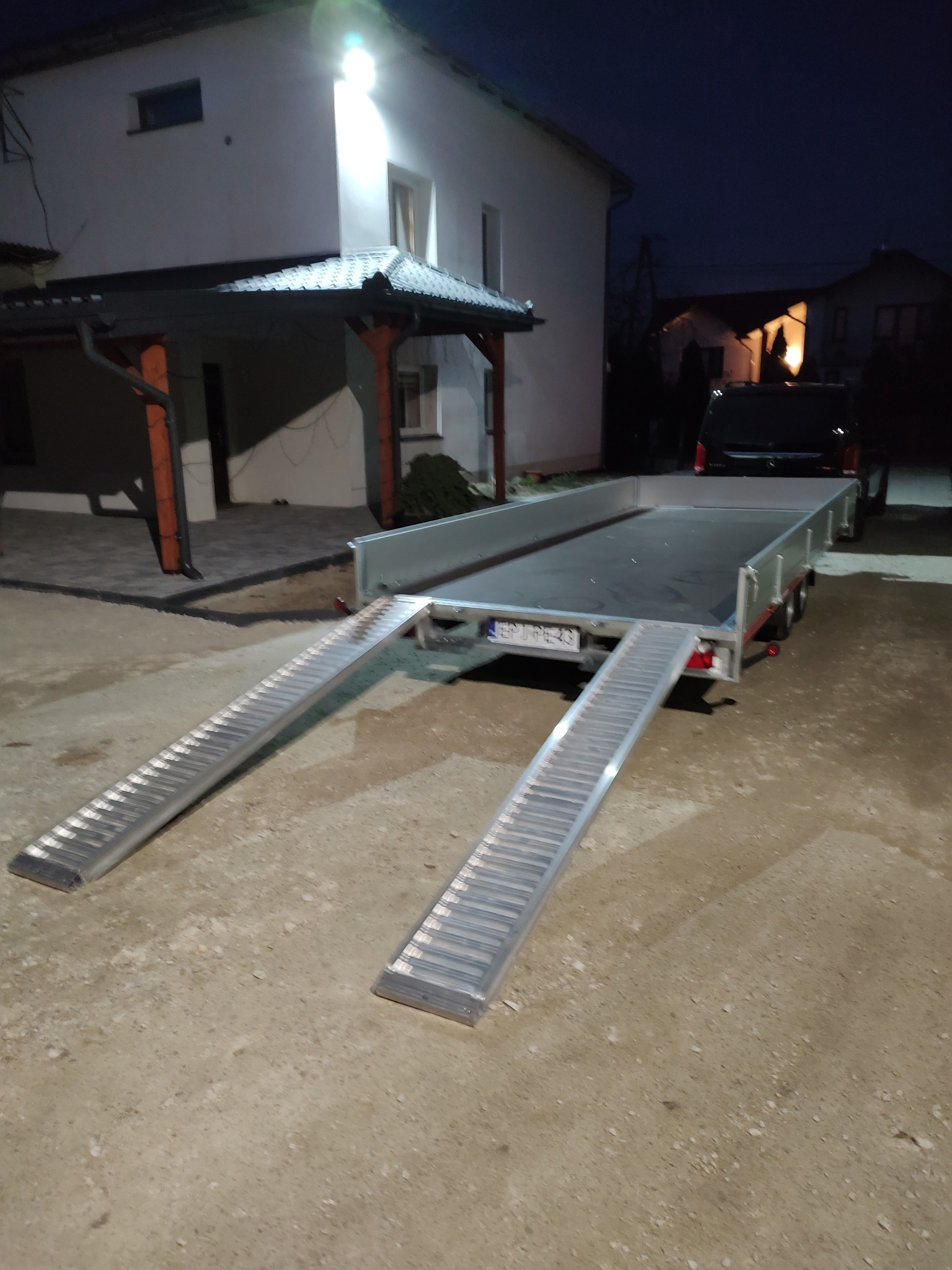 Nowa, aluminiowa przyczepa samochodowa z rozłożonymi najazdami, gotowa do transportu, na podwórku w Siemkowicach, w tle dom jednorodzinny i samochód dostawczy.