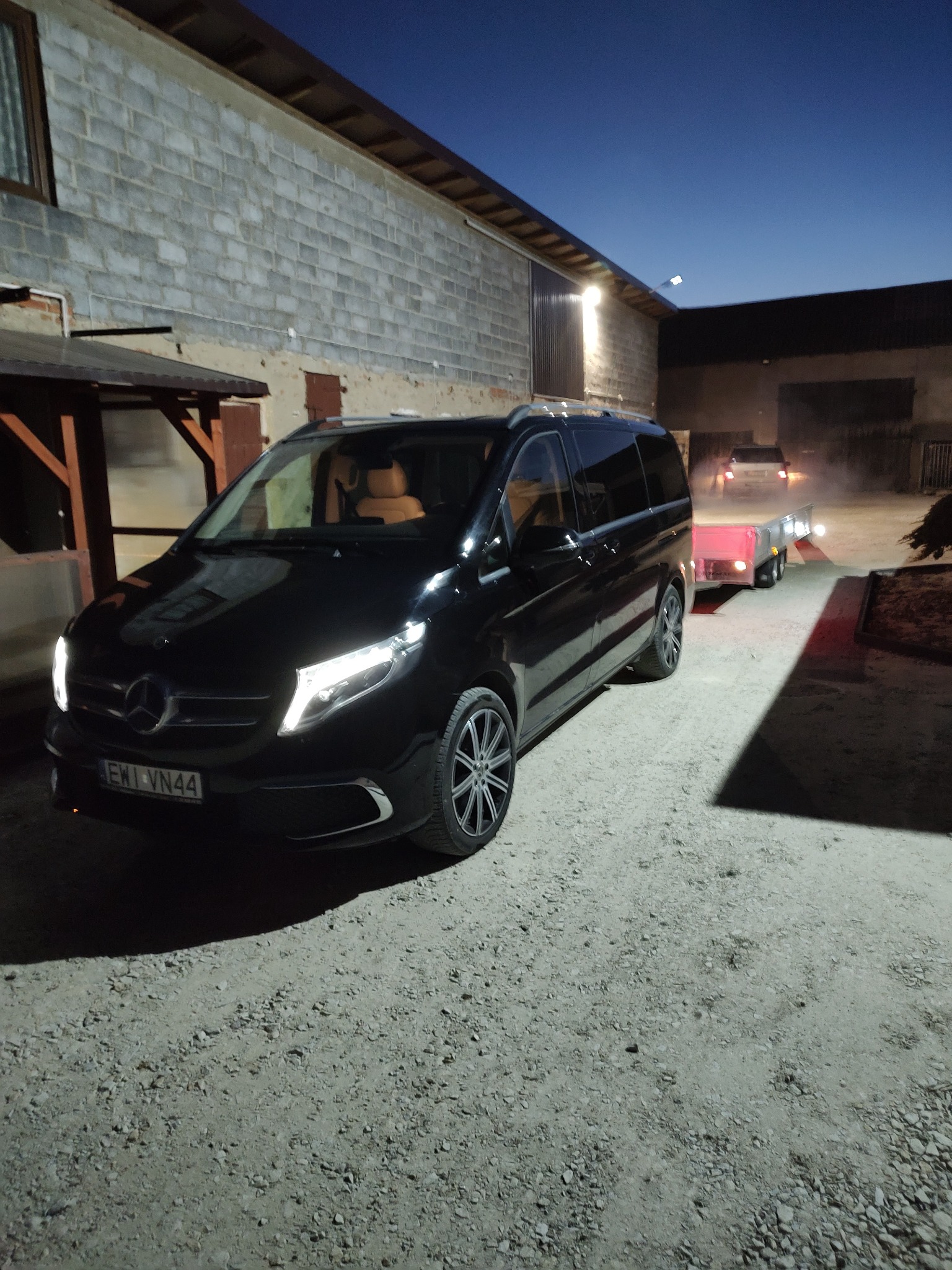 Czarny Mercedes V-klasa z przyczepą na żwirowym podłożu, oświetlony światłami, z budynkiem w tle i zmierzchem na niebie.