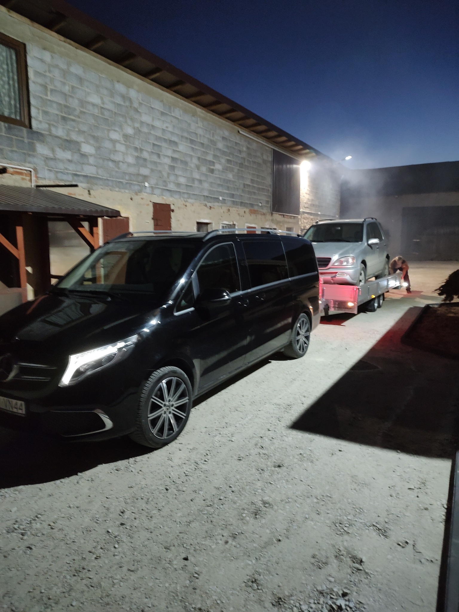 Czarny van Mercedes z włączonymi światłami LED stoi przed lawetą z załadowanym srebrnym SUV-em na tle szarego budynku gospodarczego w słabym oświetleniu nocnym.