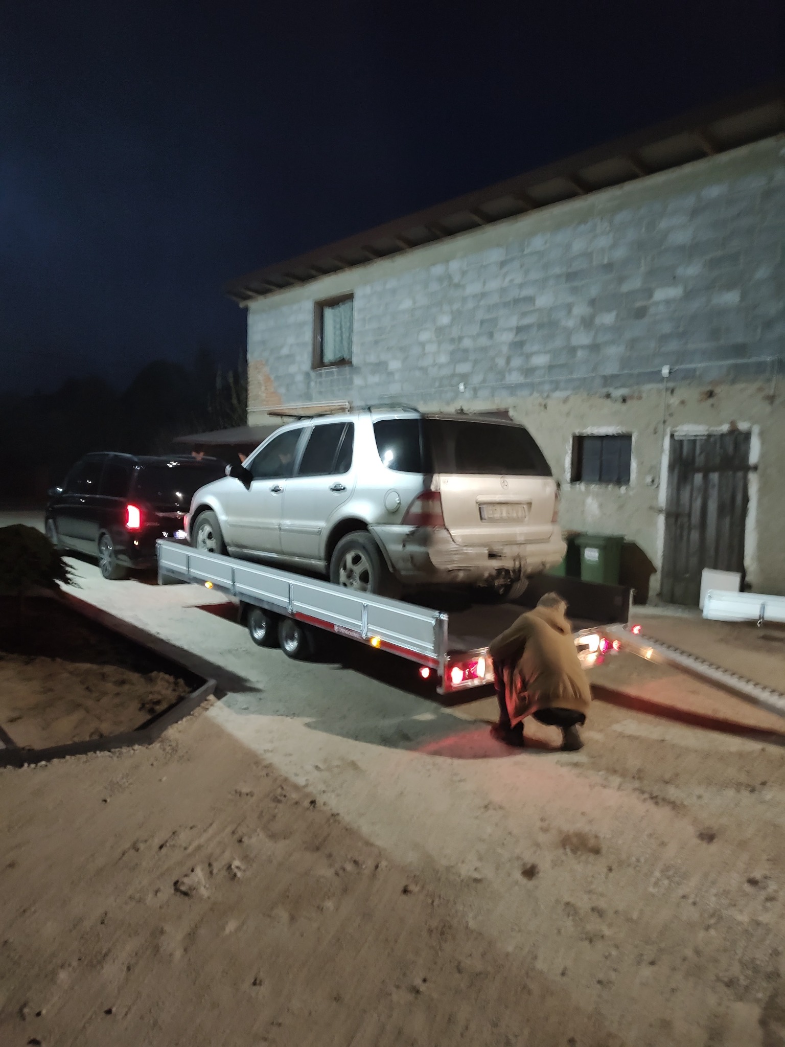 Srebrny SUV z uszkodzonym tyłem załadowany na platformę przyczepy w słabym oświetleniu, osoba kuca przy platformie, w tle ciemny budynek gospodarczy.