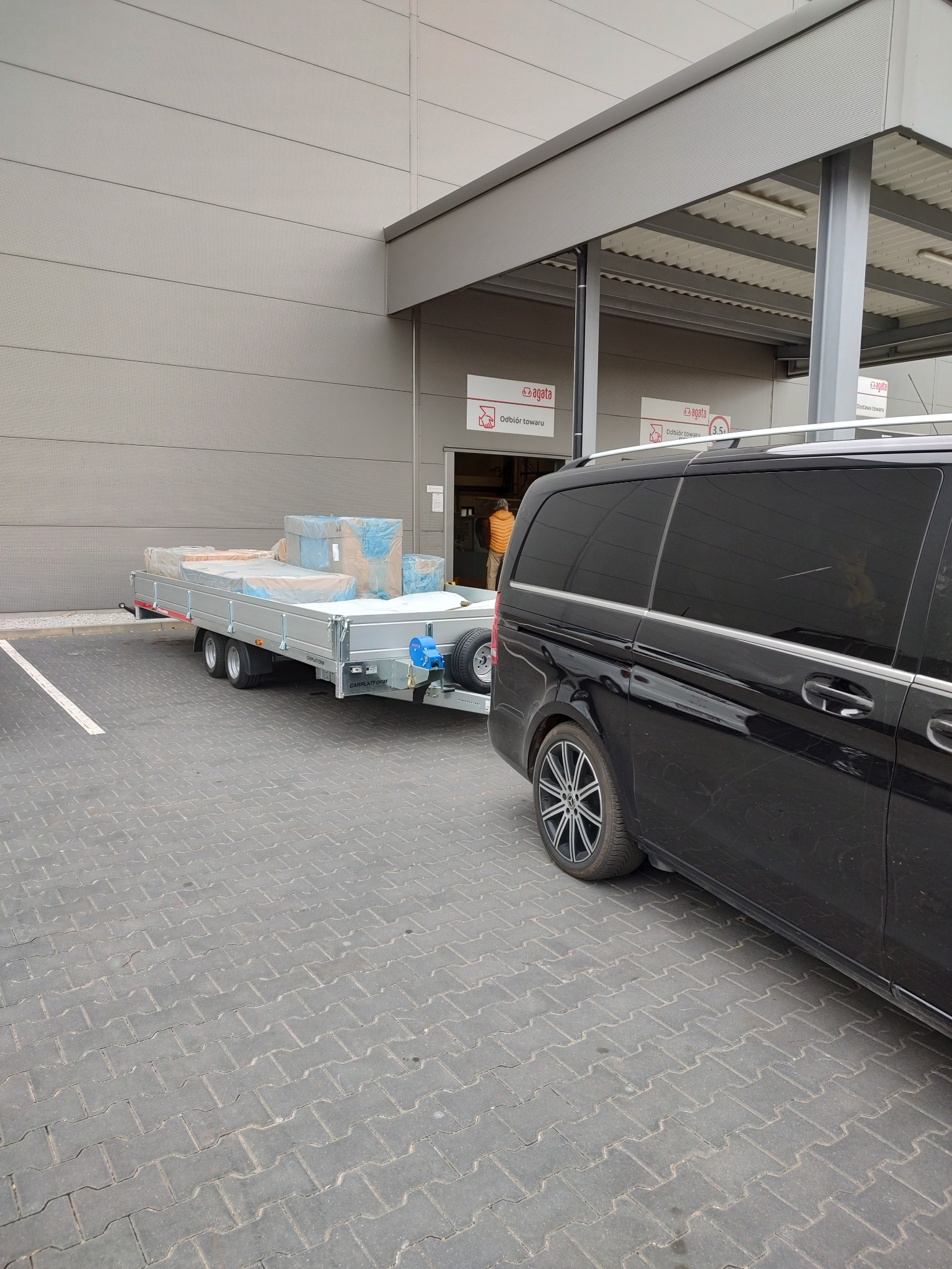 Czarny van z przyczepą platformową zaparkowany na kostce brukowej przed budynkiem Agata Meble, na przyczepie ładunek zabezpieczony folią.