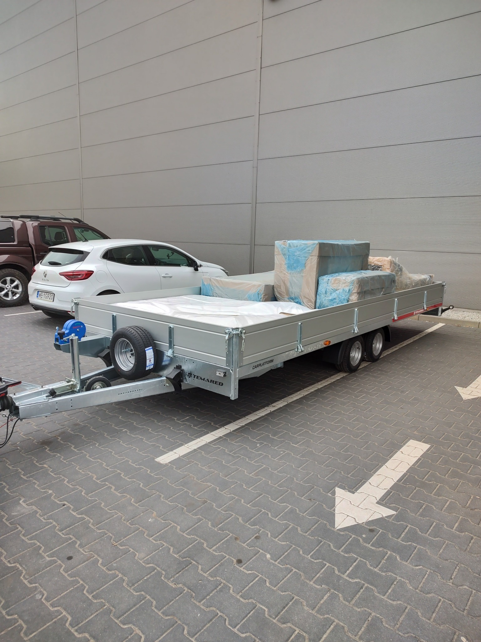 Szara przyczepa samochodowa marki Temared załadowana towarami zabezpieczonymi folią, zaparkowana na parkingu z kostki brukowej, obok zaparkowane samochody osobowe.