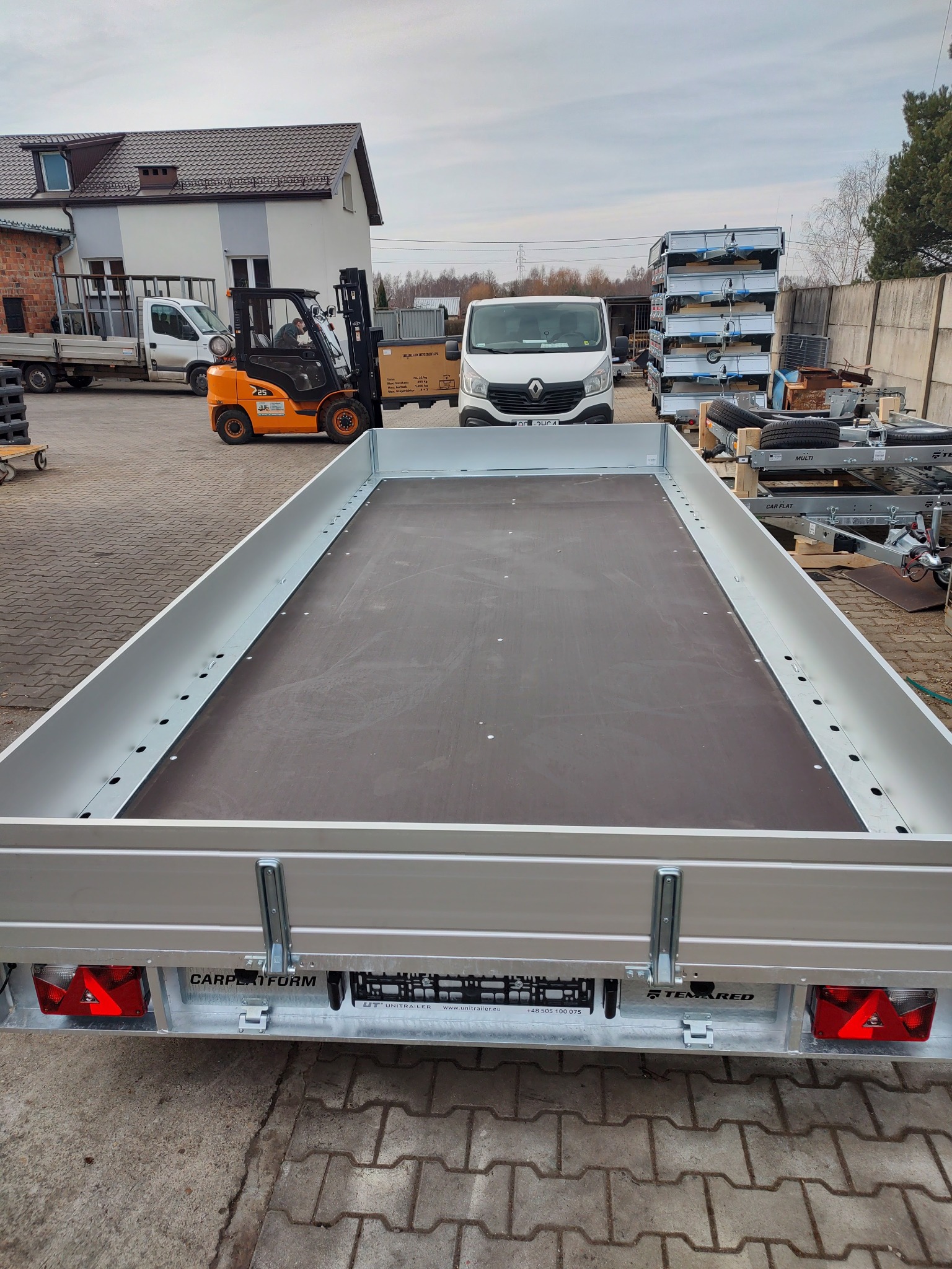 Widok z tyłu nowej, pustej przyczepy samochodowej z drewnianą podłogą, na tle budynków, wózka widłowego i dostawczego busa.