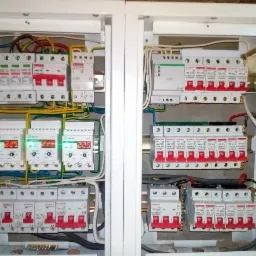 Rozdzielnica elektryczna z widocznymi bezpiecznikami, przewodami i wyświetlaczami cyfrowymi wskazującymi wartości napięcia, zamontowana w białej obudowie.